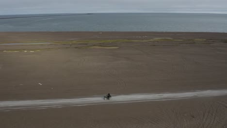 Toma-Aérea-De-Drones-De-Seguimiento-De-Un-Hombre-Solitario-En-Atv-En-La-Playa-De-Arena-Negra-Y-El-Océano-ártico-En-El-Punto-Más-Septentrional-Del-ártico-Estados-Unidos-Cerca-De-Barrow-Alaska