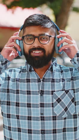 Portrait-of-happy-smiling-Indian-man-listening-music-taking-off-headphones-looking-at-camera-outdoor