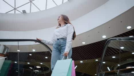 young woman holding shopping bags steps onto descending escalator in modern shopping mall, placing hand on rail while looking into distance, surrounded by bright lights, stylish decor