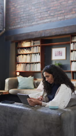 vertical video of biracial woman sitting on sofa and using tablet at home, slow motion