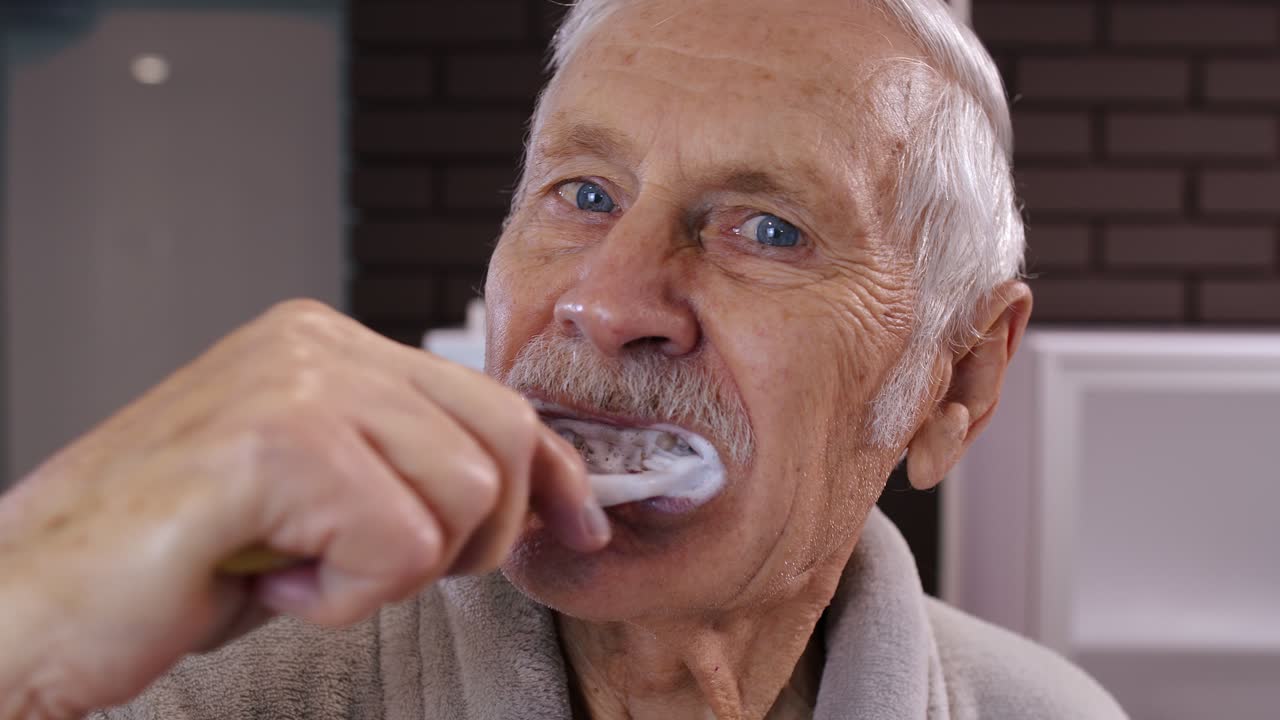 Senior man grandfather in bathrobe brushing teeth, looking into mirror. Morning hygiene at bathroom