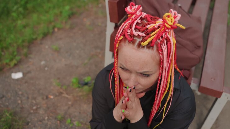 Donna bianca con dreadlocks rosa che implora silenziosamente, mani giunte vicino al mento, seduta su una panchina rossa in un piccolo parco, fronte tesa e occhi speranzosi, sfondo urbano, forte tensione emotiva