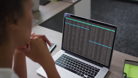 mujer biracial sentada en el escritorio viendo el procesamiento de datos de codificación en la pantalla de la computadora portátil