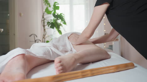 massage therapist wearing black uniform focuses on massaging client thigh with firm palm technique while background shows soft daylight, indoor plant, and decoration blurred spa room setting