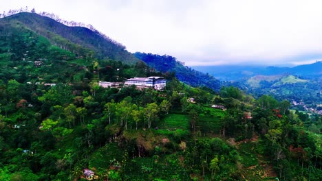 a lush sri lankan hillside features a large building surrounded by green slopes, scattered village houses, and breathtaking views of layered tropical mountains under a soft cloudy sky.