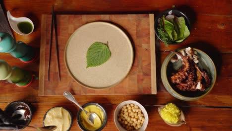 Table-top-down-view-of-professional-fine-dinning-chef-plating-delicious-grilled-octopus-dish,-by-placing-decorative-peppermint-leaf-on-empty-plate,-a-scoop-of-mashed-potato-and-chargrilled-tentacles