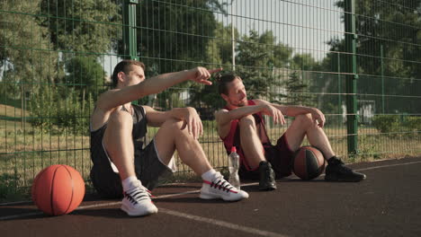 dos felices jugadores de baloncesto charlando sentados y apoyados contra una cerca de metal en una cancha de baloncesto al aire libre