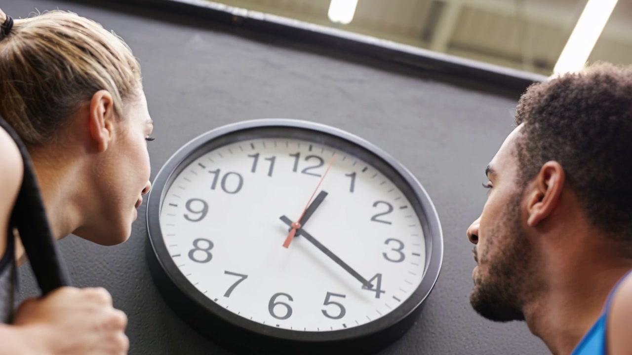 Donna in abbigliamento sportivo che controlla i compagni di palestra in pausa tra le serie, tenendo un asciugamano, vicino all'orologio a parete della palestra.