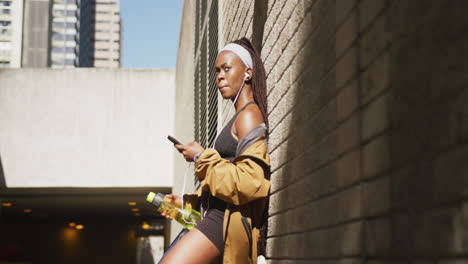 mujer afroamericana haciendo ejercicio al aire libre usando auriculares usando un teléfono inteligente en la ciudad