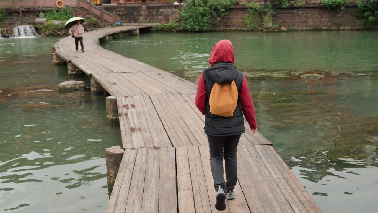 Penyeberangan Jembatan Kayu Tepi Danau oleh Pelancong, Backpacker Berhoodie Merah Berjalan Menjauh dari Kamera di Sepanjang Papan Lapuk, Air Hijau Tenang Memantulkan Langit Mendung, Pejalan Kaki di Kejauhan Berhenti di Bawah Payung