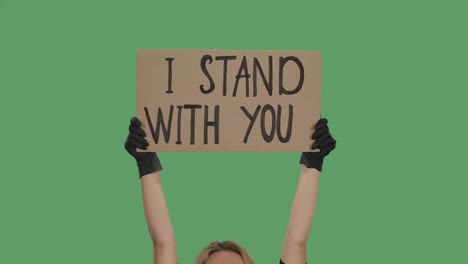 blond woman holding over head a poster from a cardboard box with the words i stand with you with both hands. anti black racism. isolated a green screen, chroma key. close up. slow motion