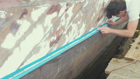 white male placing blue masking tape on wooden liveaboard hull before painting