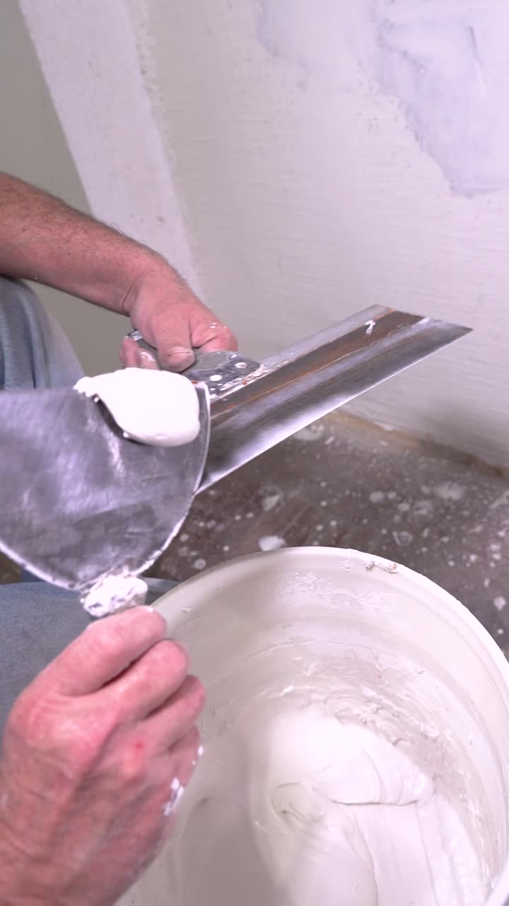 A close-up of a person's hand mixing white joint compound or plaster in a large bucket using a wide trowel for home renovation