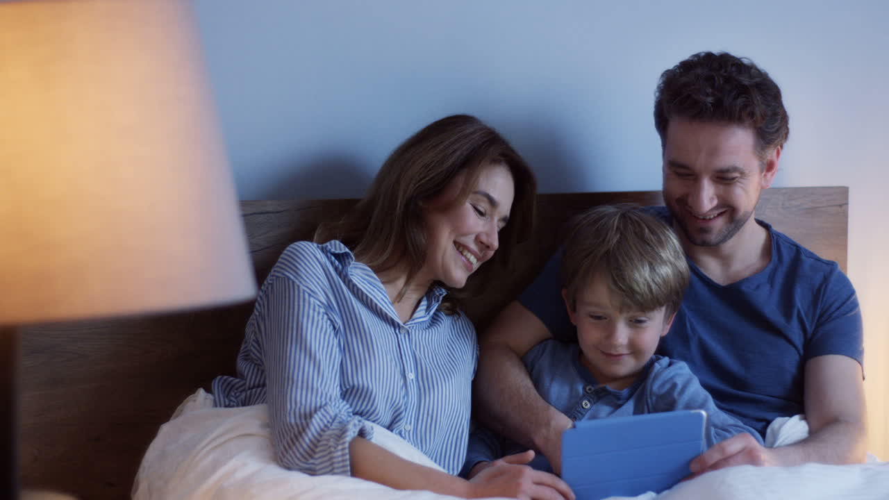 Caucasian parents sitting on the bed with their little cute son, watching something on the tablet and talking at night