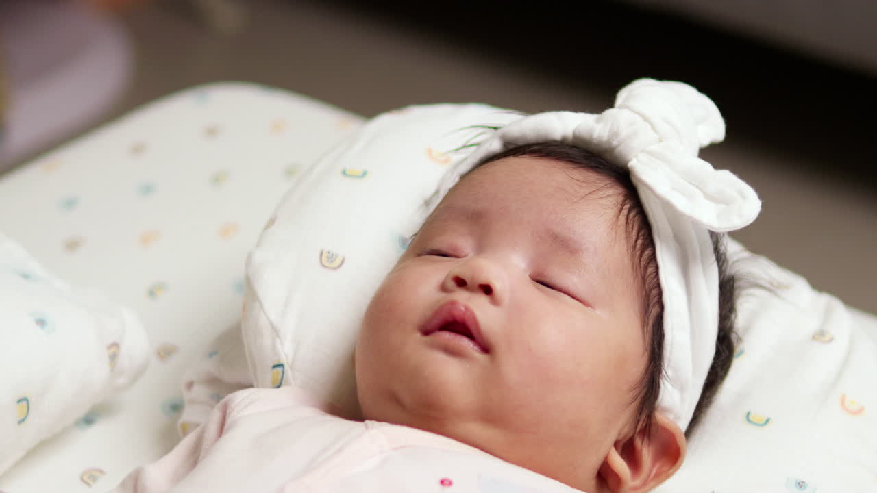 Puzzled baby in pink outfit feeling drowsy on the bed with a white bow on her head. Her chubby cheeks and round eyes make this moment irresistibly cute