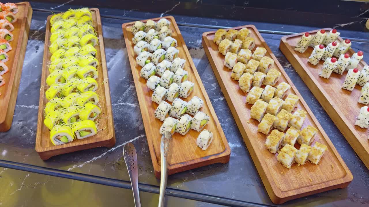 Camera pans across assorted maki sushi with salmon, avocado and sesame on wooden trays