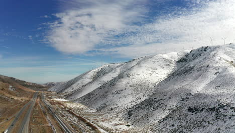 Snowy-mountains-by-highway-with-blue-sky-and-clouds,-Aerial