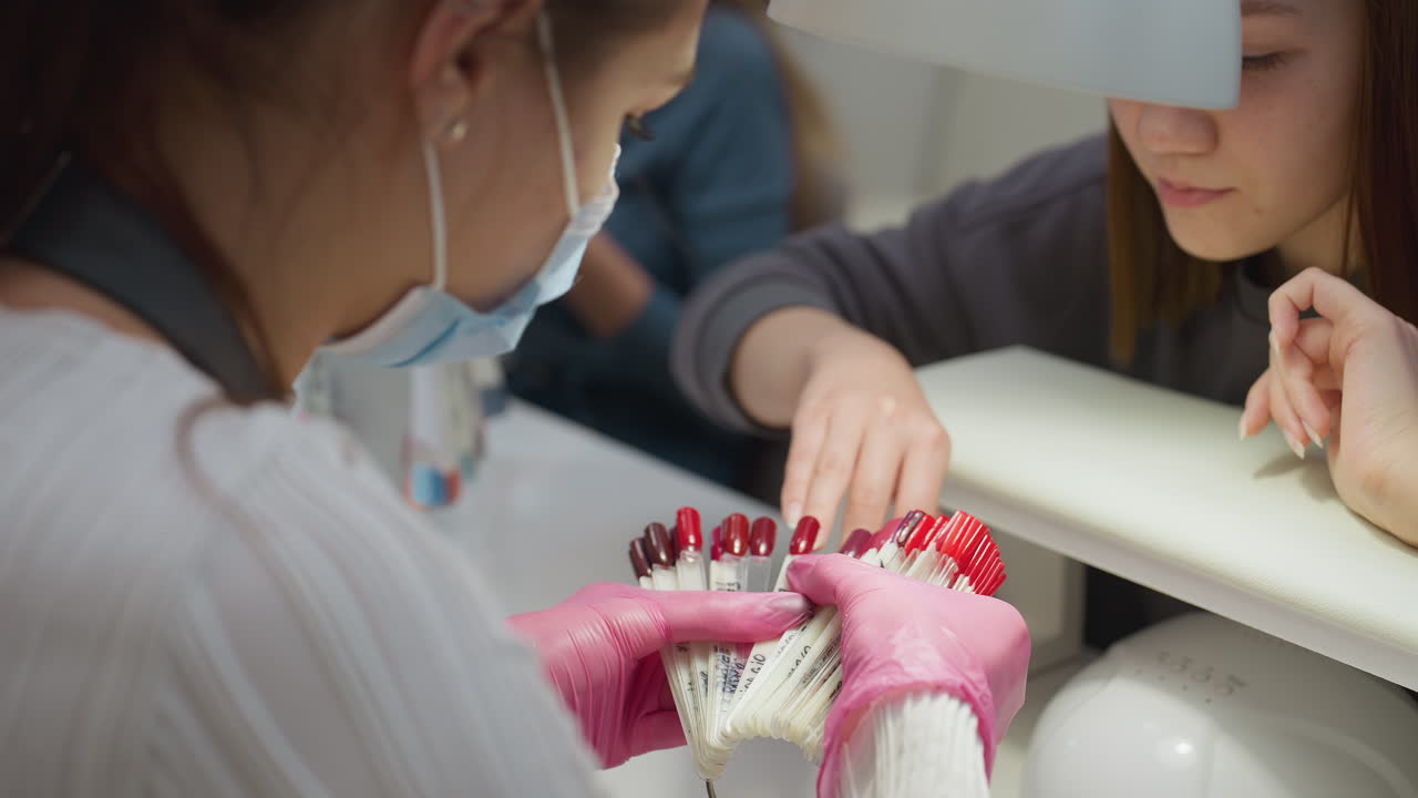 Nail technician wearing pink gloves and face mask holds up red polish color samples while customer attentively evaluates options under bright salon lamp during personalized manicure consultation