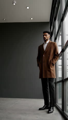 stylish man in a brown coat standing by large windows in a modern building