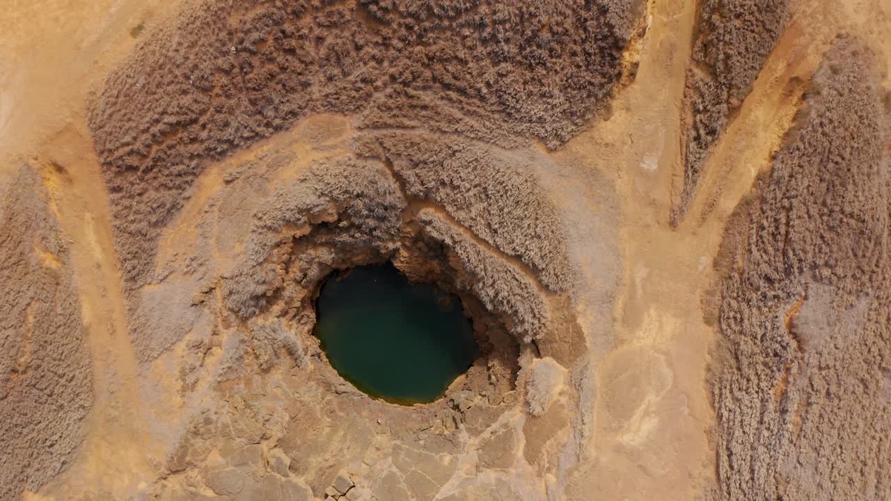 From directly above, a circular collapse pit holds emerald water, its rock rim scalloped by erosion while wheel tracks converge across the arid plain, suggesting geology, fragility, and solitude