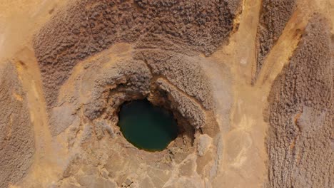 from directly above, a circular collapse pit holds emerald water, its rock rim scalloped by erosion while wheel tracks converge across the arid plain, suggesting geology, fragility, and solitude