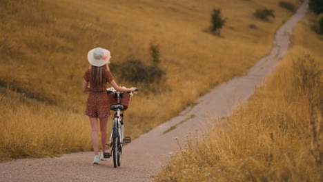 Rückansicht-In-Zeitlupe:-Eine-Schöne-Blondine-In-Einem-Kleid-Mit-Blumen-In-Einem-Korb-Und-Einem-Retro-Fahrrad-Geht-Auf-Der-Sommerwiese-Die-Straße-Entlang,-Schaut-Sich-Um-Und-Lächelt-Mit-Einem-Gefühl-Der-Freiheit.