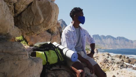 African-american-man-wearing-face-mask-hiking-in-mountain-countryside-sitting-on-a-rock