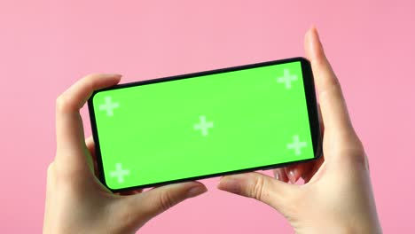 close-up of female hands holding smartphone with blank green screen showing copy space for app or website design, on pink background , mockup.