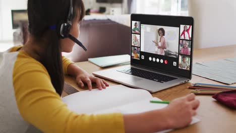 Schoolgirl-using-laptop-for-online-lesson-at-home,-with-diverse-teacher-and-class-on-screen