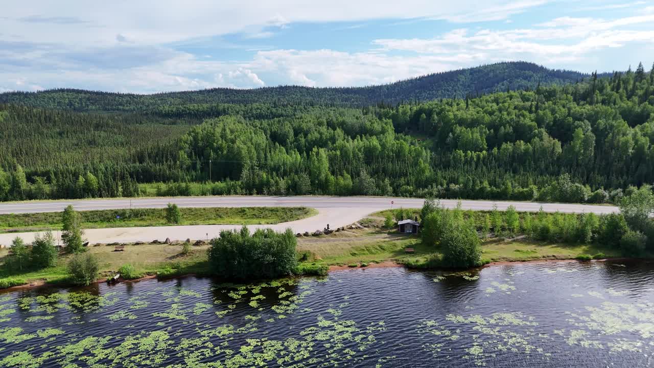 Scenic drive past a pristine lake surrounded by lush green forests under a clear blue sky in Alaska, perfect for travel adventures and outdoor exploration