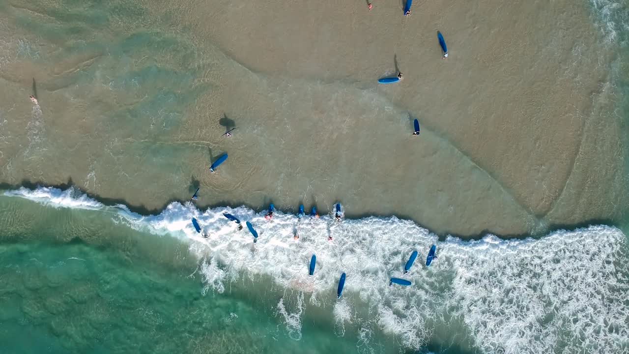 disparo aéreo de drones mirando hacia abajo a un gran grupo de personas aprendiendo a surfear en la playa
