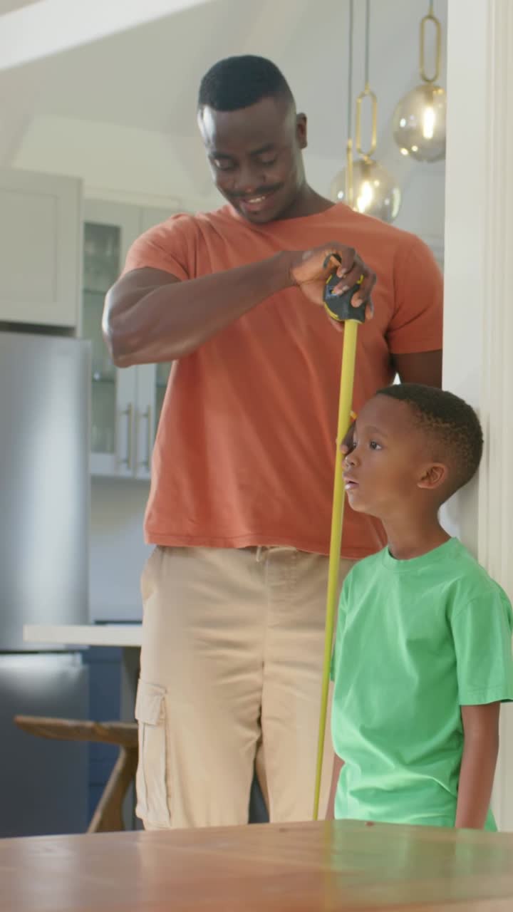 Vertical video: Pulling tape measure from case, dad measuring boy in tee checking growth in kitchen