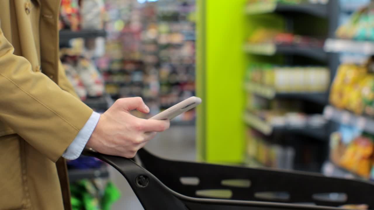 Close up of man hand holding smartphone and srolling screen in supermarket. Man using phone while walking and pushing shopping cart in supermarket.
