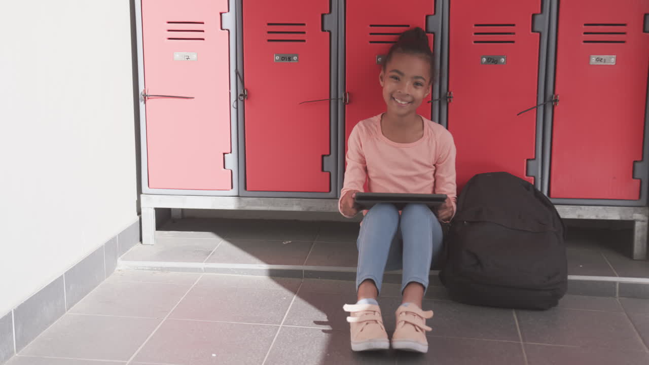 Sitting by lockers, African American girl using tablet and smiling in school hallway, copy space