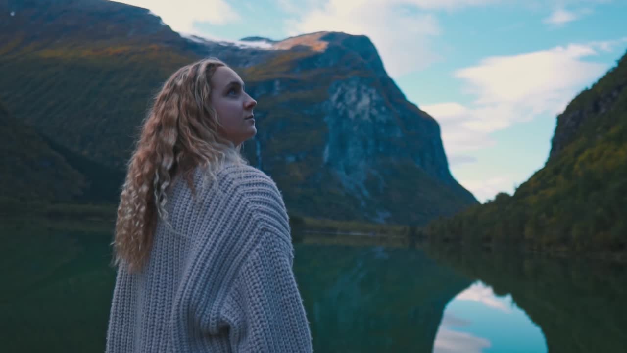 Slow motion shot of a blonde girl, looking from the left to her right at the sky, large mountains and sea in the background. Norway