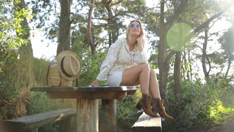 woman leisurely sitting on a table in forest landscape a woman is seated on a wooden table surrounded by trees in the forest with lush green grass and plants all around she is leisurely enjoying the scenic landscape wearing eyewear