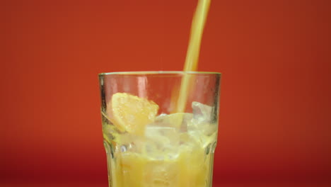 jugo de naranja fresco vertido y derramado en un vaso alto lleno de bloques de hielo y naranjas, aislado en un fondo naranja