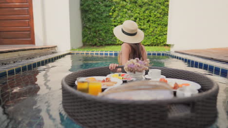 asian woman in mustard color bathing suit pulls on her floating tray of breakfast inside a pool