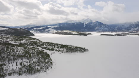 Aerial-rotating-right-to-reveal-icy,-frozen-lake-surrounded-by-snow-and-green-pine-trees-with-large-snowy-ski-town-mountains-in-the-background-near-Silverthorne-and-Frisco-Colorado