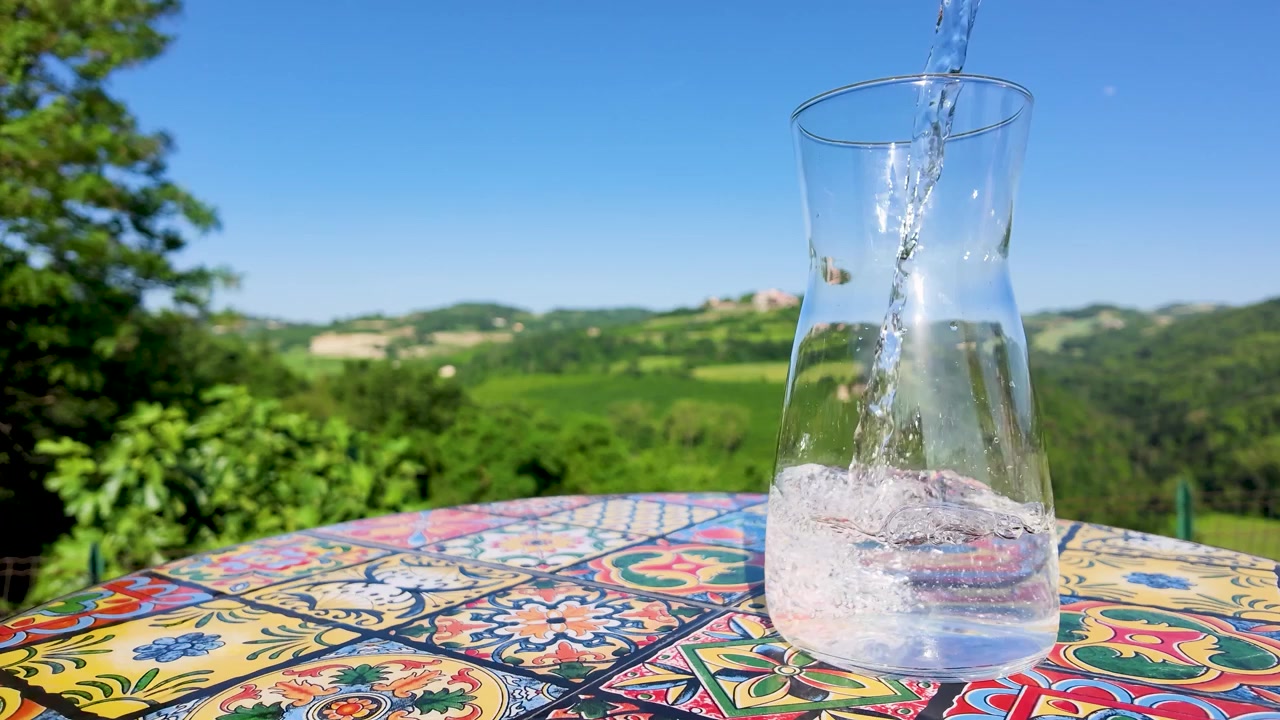 Versamento al rallentatore di acqua frizzante in una caraffa di vetro su una terrazza mediterranea.