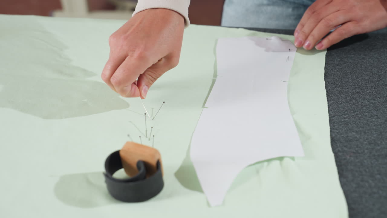 Clothing designer carefully removes pins used to secure white paper pattern over elastic light green textile while preparing garment layout on professional sewing workspace in bright creative studio