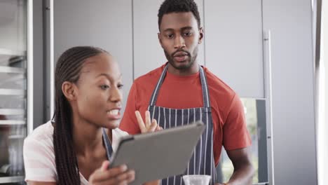 happy african american couple in aprons baking together using tablet in kitchen, slow motion