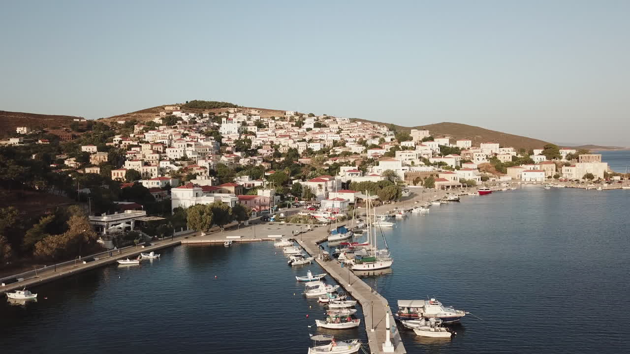 Greece, Ionusses Island, Drone Footage of the harbor on a sunny day with calm seas.Smaller boats inside the pier and sailboats outside, camera turns around and moves towards the town with white houses