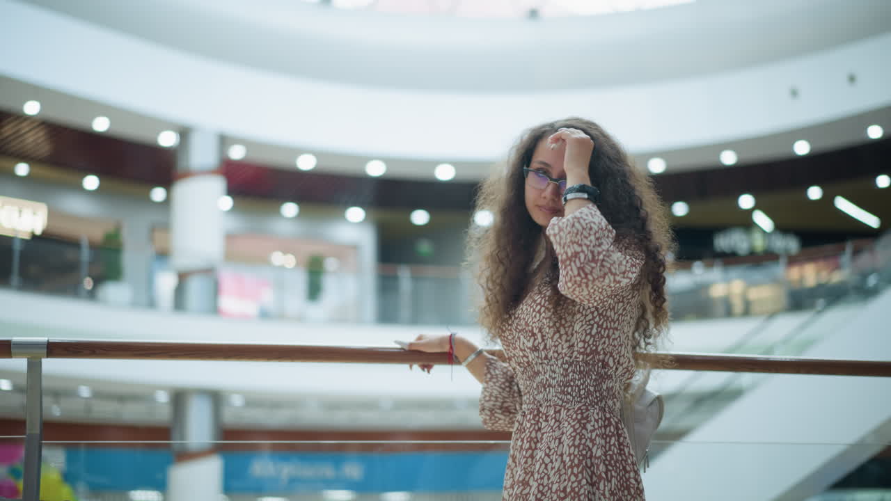 mujer en vestido vintage posa junto a una barandilla de hierro en un amplio centro comercial, ajustando su cabello con una expresión reflexiva, el fondo presenta compradores borrosos, iluminación suave y detalles arquitectónicos