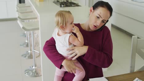 caucasian mother holding her baby and talking on smartphone at home