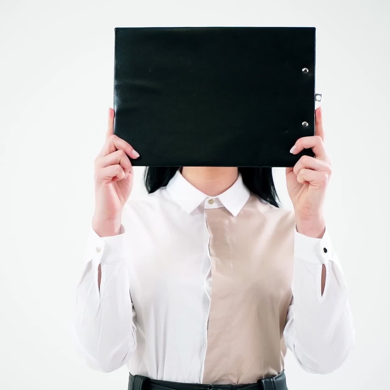 Black folder hides woman face. Attractive secretary opens her face hidden with a folder and smiles isolated on white wall. Businesswoman with positive emotions.