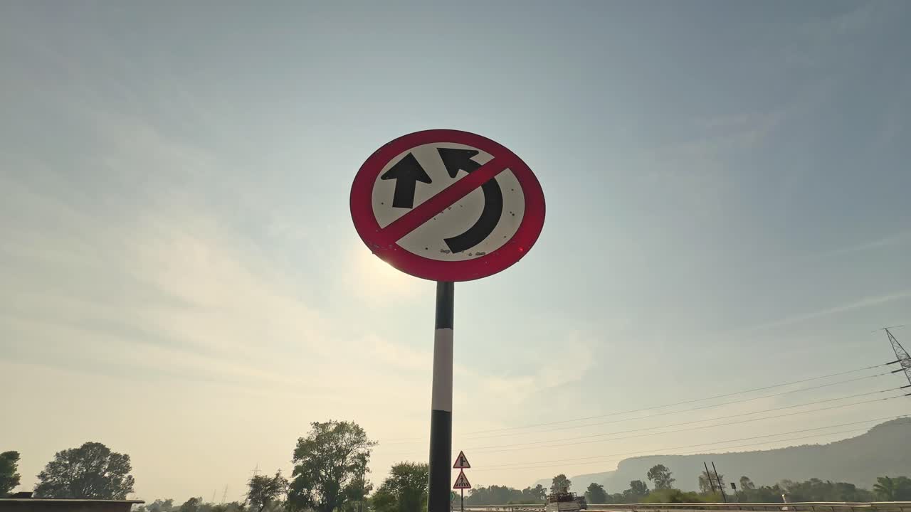 Static shot of a no U-turn road sign mounted on a pole, framed against clear sky with distant trees and highway elements in soft daylight