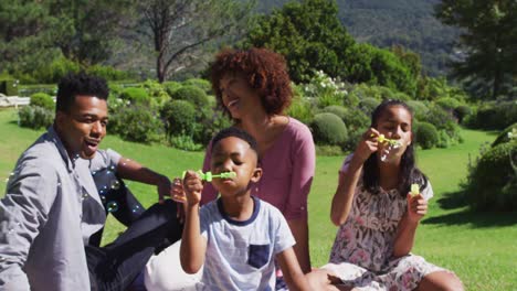 happy african american parents, daughter and son sitting outdoors on blanket, blowing bubbles