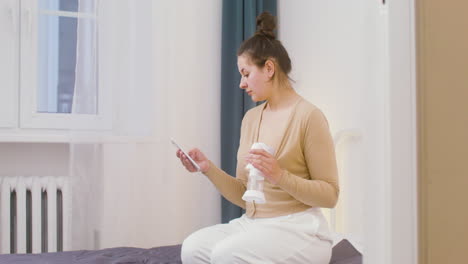 Young-Mother-Using-Breast-Pump-And-Scrolling-On-Mobile-Phone-While-Sitting-On-The-Bed-At-Home-1