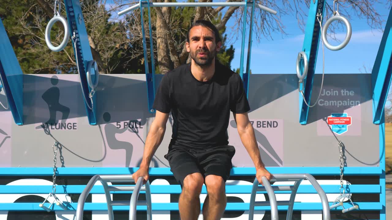Hispanic Man with beard Does Hanging Leg Raises on Park Fitness Course, medium shot